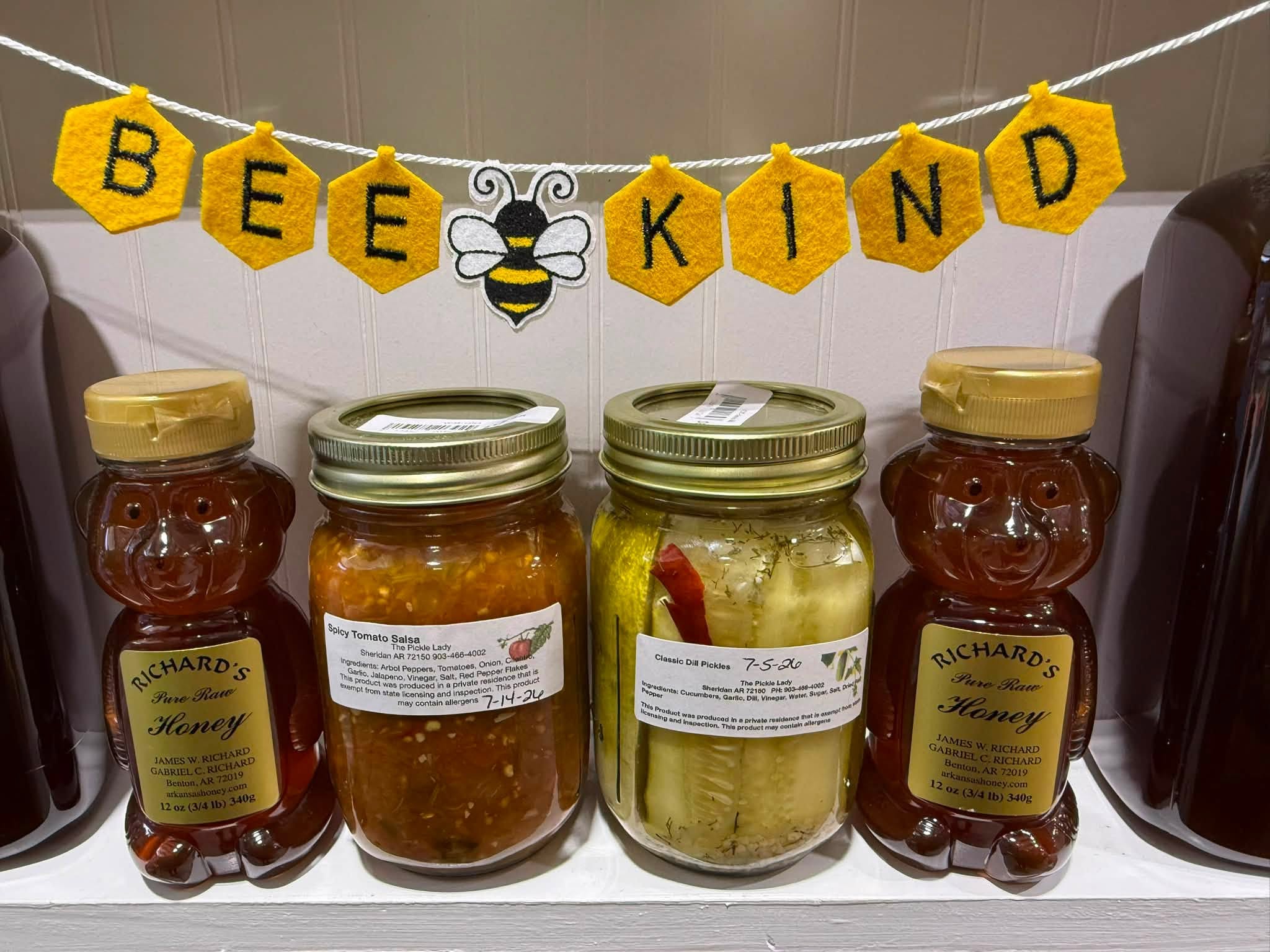 Local honey, salsa, and pickles on a shelf with a Bee Kind banner